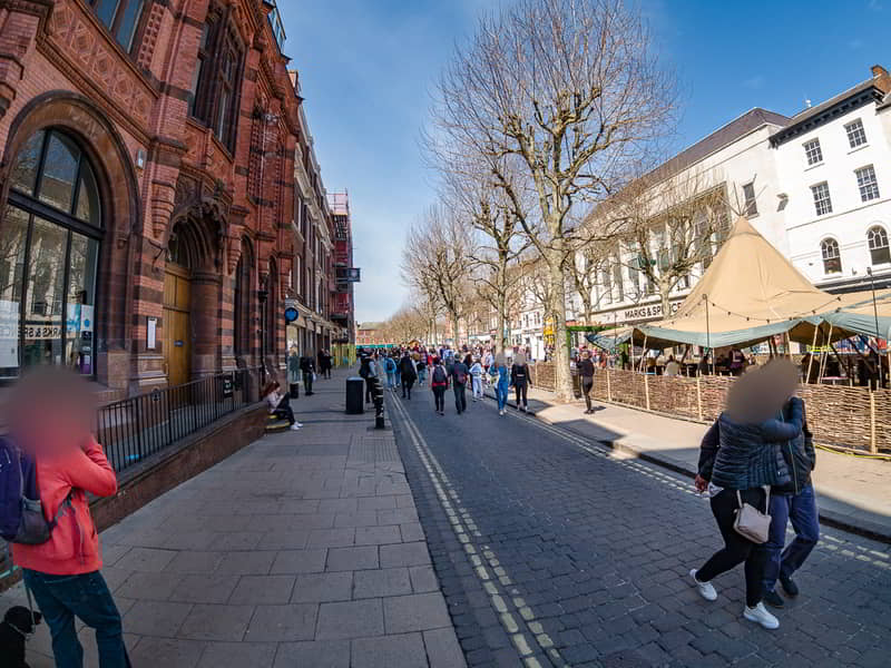 Street view image of Parliament Street in York on a bright sunny day. People are casually walking down the pedestrian area. The left side of the photo shows the ornate brick facade of a historic building, while the right side displays open-air market stalls under canvas umbrellas. The faces of individuals have been blurred for privacy.