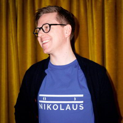 Nikolaus wears a t-shirt with his company logo on it, and glasses and a cardigan